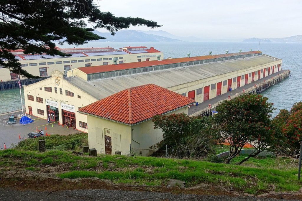 View from the hillside of the Festival Pavilion, a long cream-coloured building with red doors and red roofs beside San Francisco Bay.