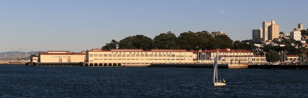 Wide view across San Francisco Bay of the long cream-coloured Fort Mason Center buildings, with the words Fort Mason Center on the roofline, a sailboat in the foreground and city buildings on the hill behind.